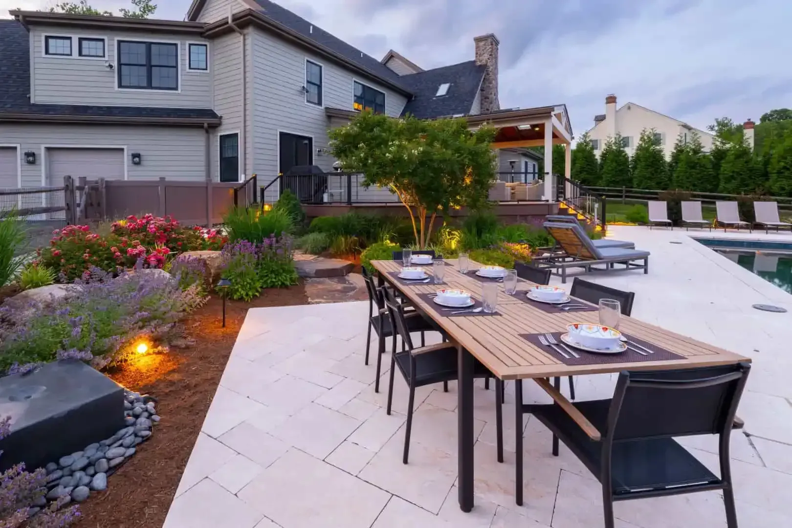 Backyard patio with outdoor dining table, black chairs, and landscaped garden beside a house at dusk