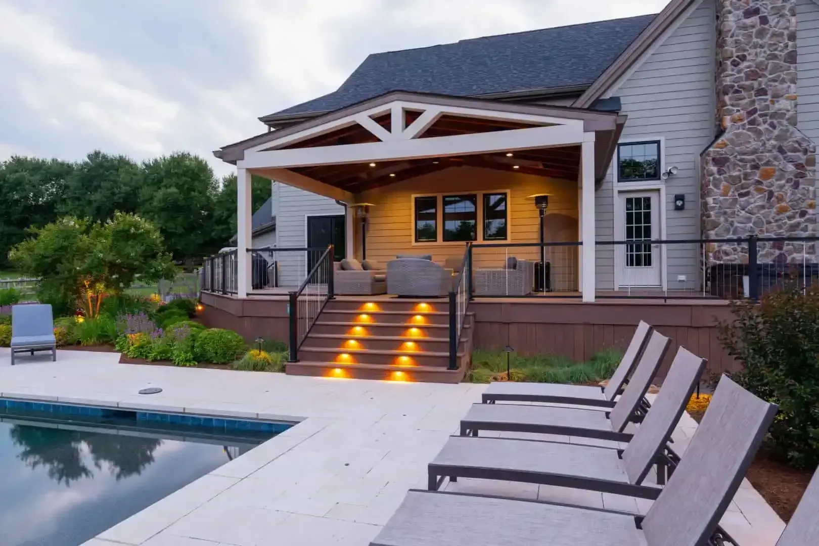 Backyard pool and patio with lit steps leading to a covered porch at dusk