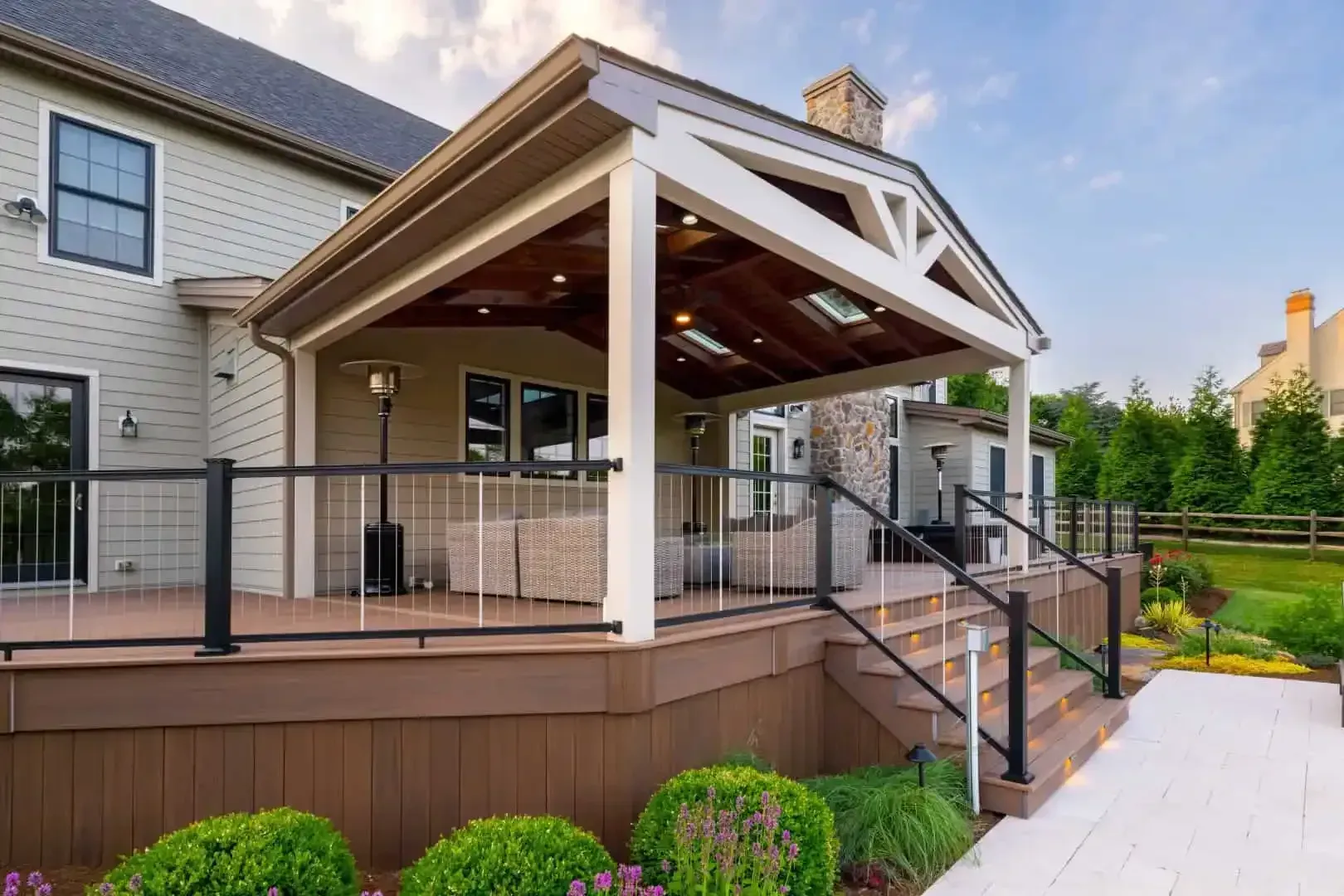 Covered wooden deck with black railing and stairs beside a beige house and landscaped yard