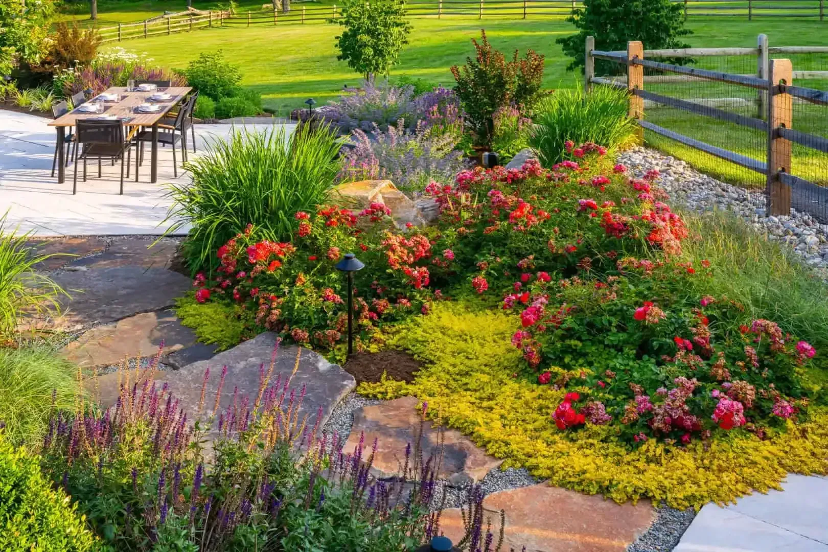 Garden patio with table and chairs beside colorful flower beds and a wooden fence in bright sunlight