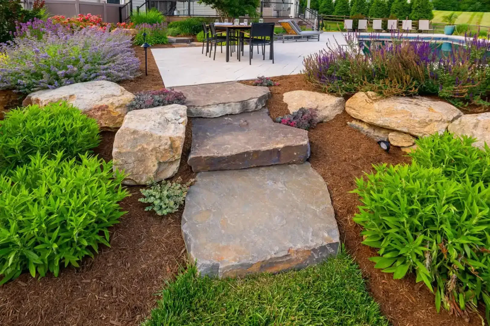 Stone garden steps leading to a patio, bordered by green shrubs and purple flowers