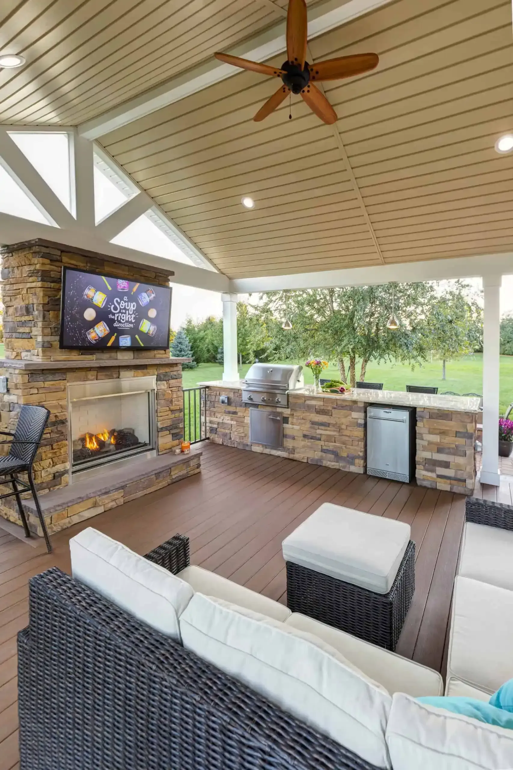 Covered outdoor patio with stone fireplace, wicker sofa, ceiling fan, and view of a grassy yard