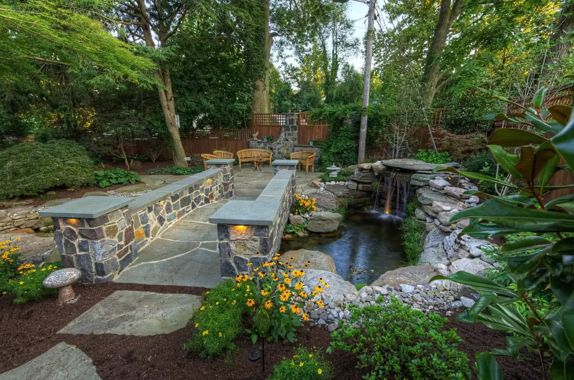 Stone patio with a small pond and waterfall, surrounded by lush garden greenery and flowers