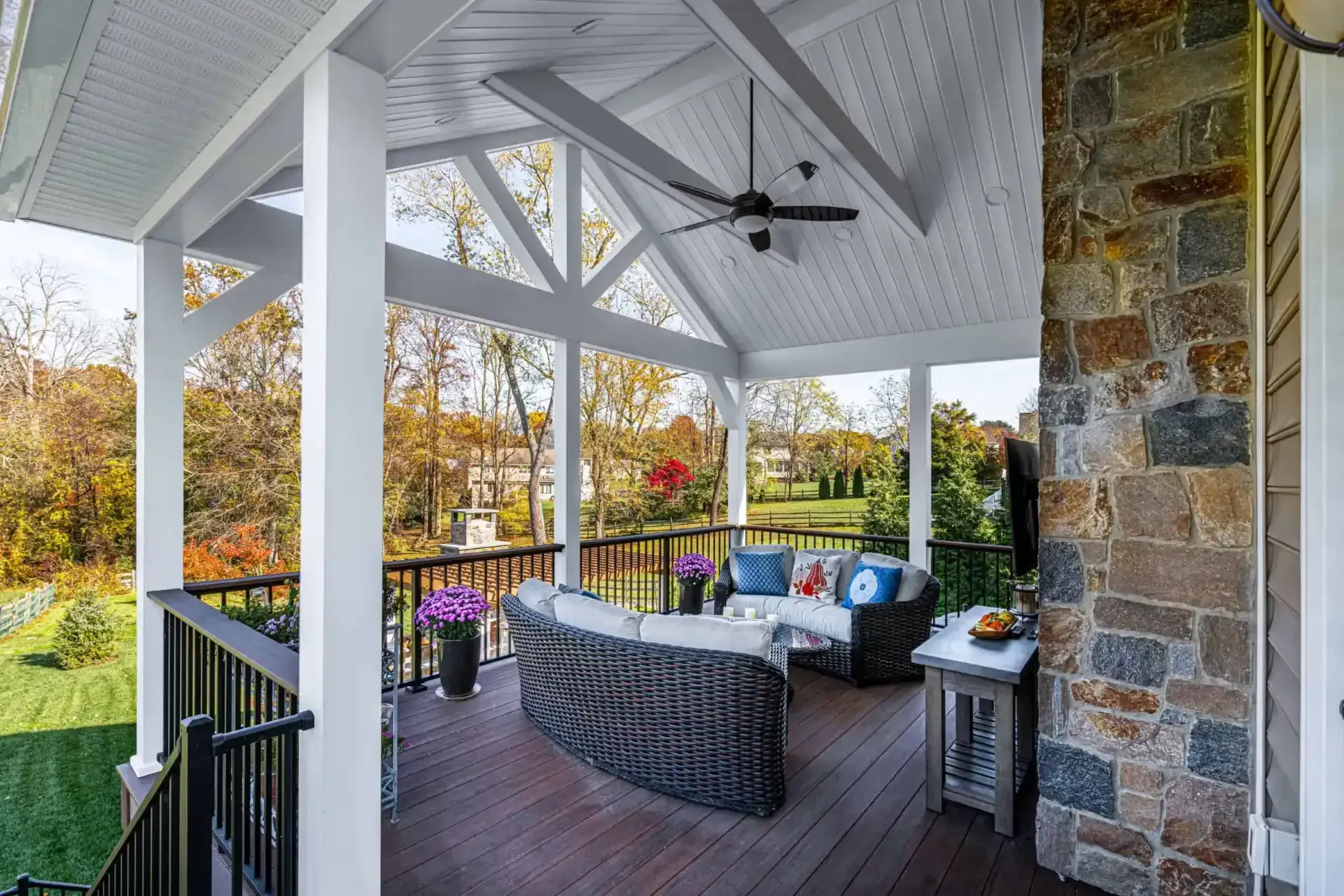 Covered porch with wicker seating, stone wall, white beams, and autumn trees beyond the railing