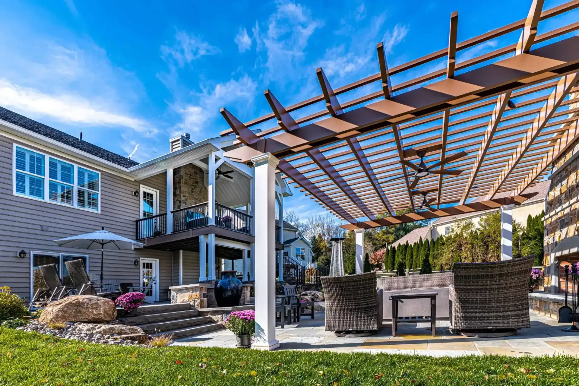 Backyard patio with pergola, outdoor seating, stone fireplace, and a two-story house under a blue sky