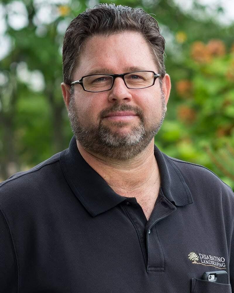 A man in glasses and black polo shirt standing outdoors with greenery in the background