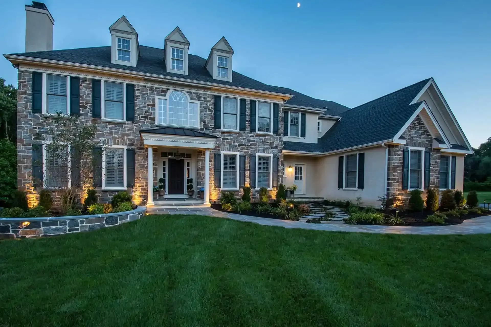 Large two-story stone house with lit exterior, white trim, and a green lawn at dusk