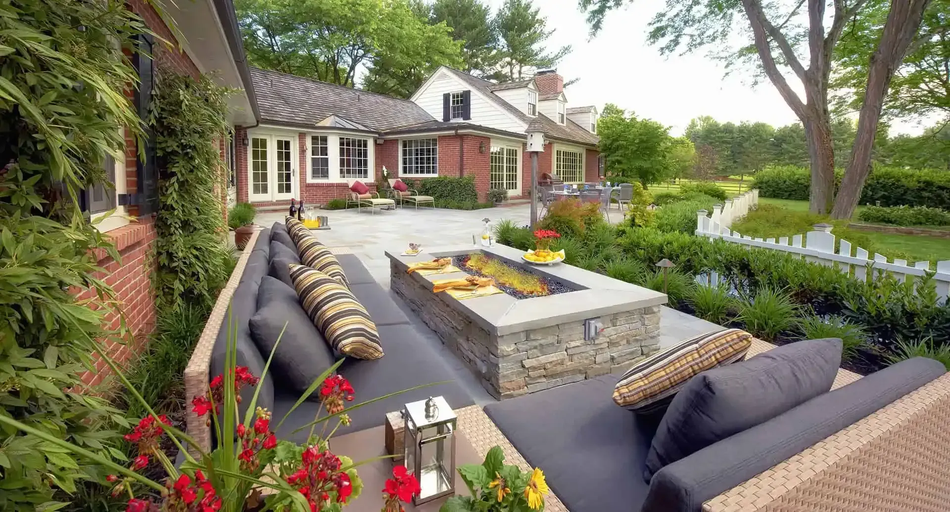 Backyard patio with fire pit, sectional seating, grill, and a red brick house surrounded by lush greenery