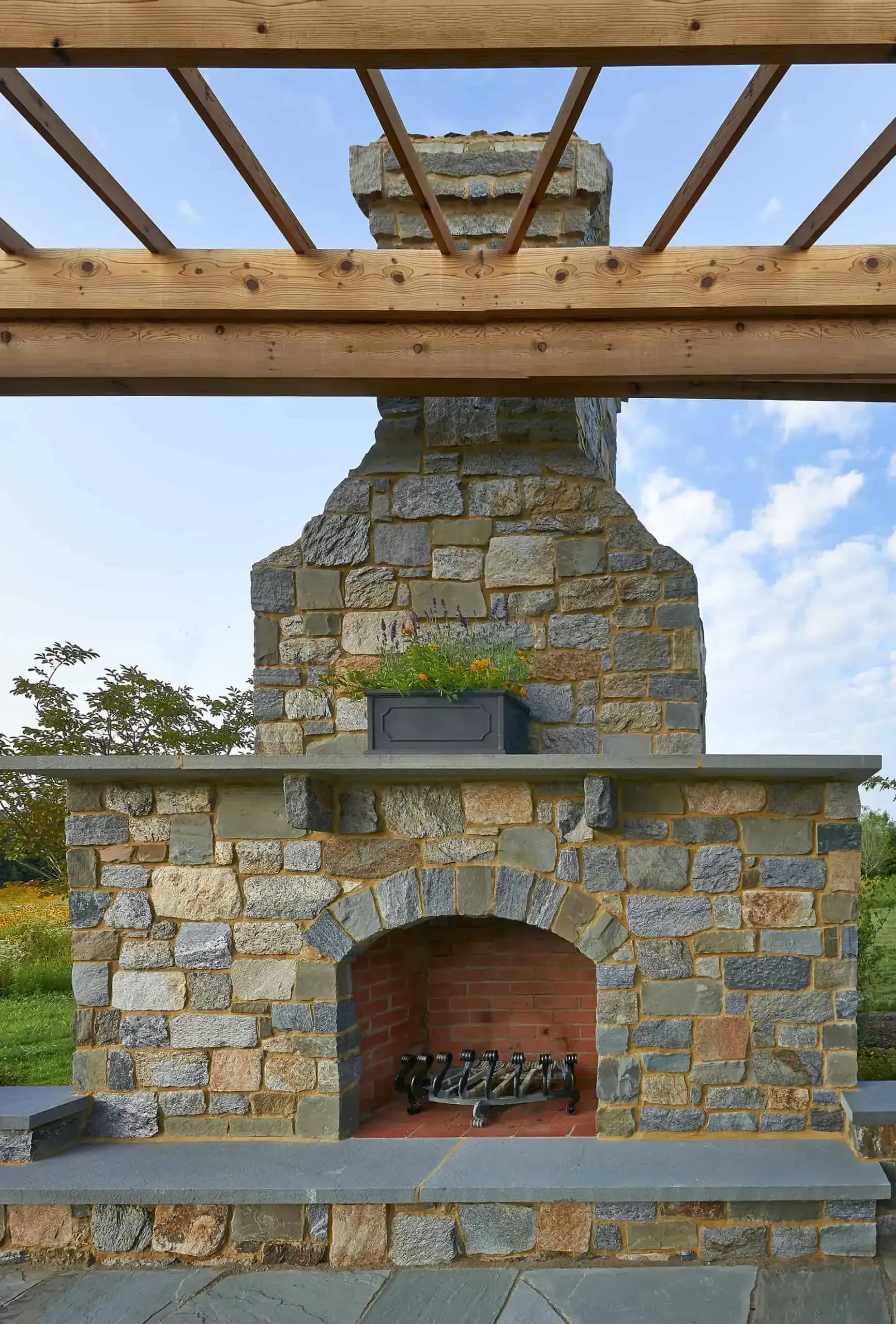 Stone outdoor fireplace with an arched firebox under a wooden pergola, set against a blue sky.