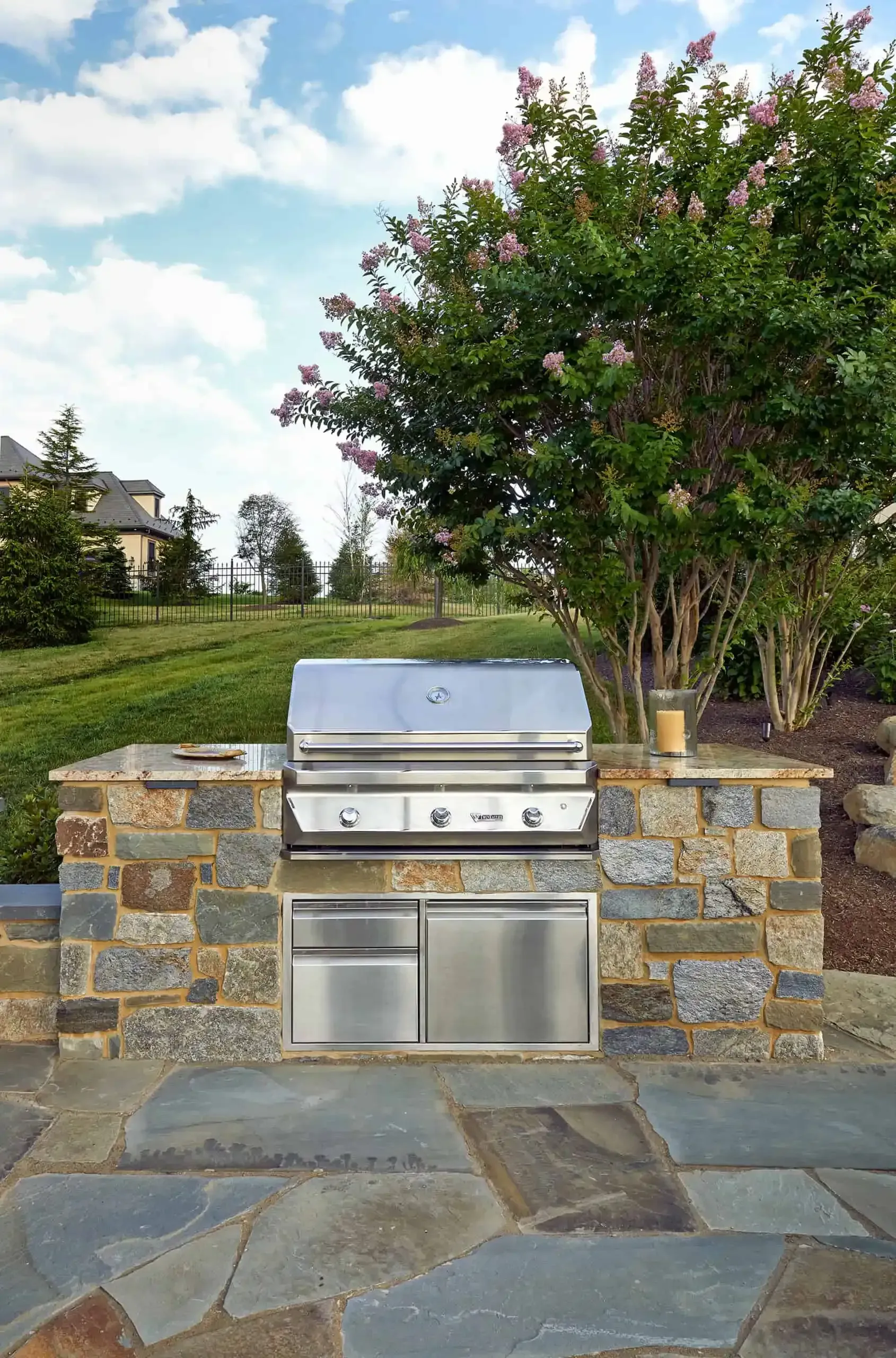 Outdoor stone patio with built-in stainless steel grill and low stone walls beside a tree and lawn.