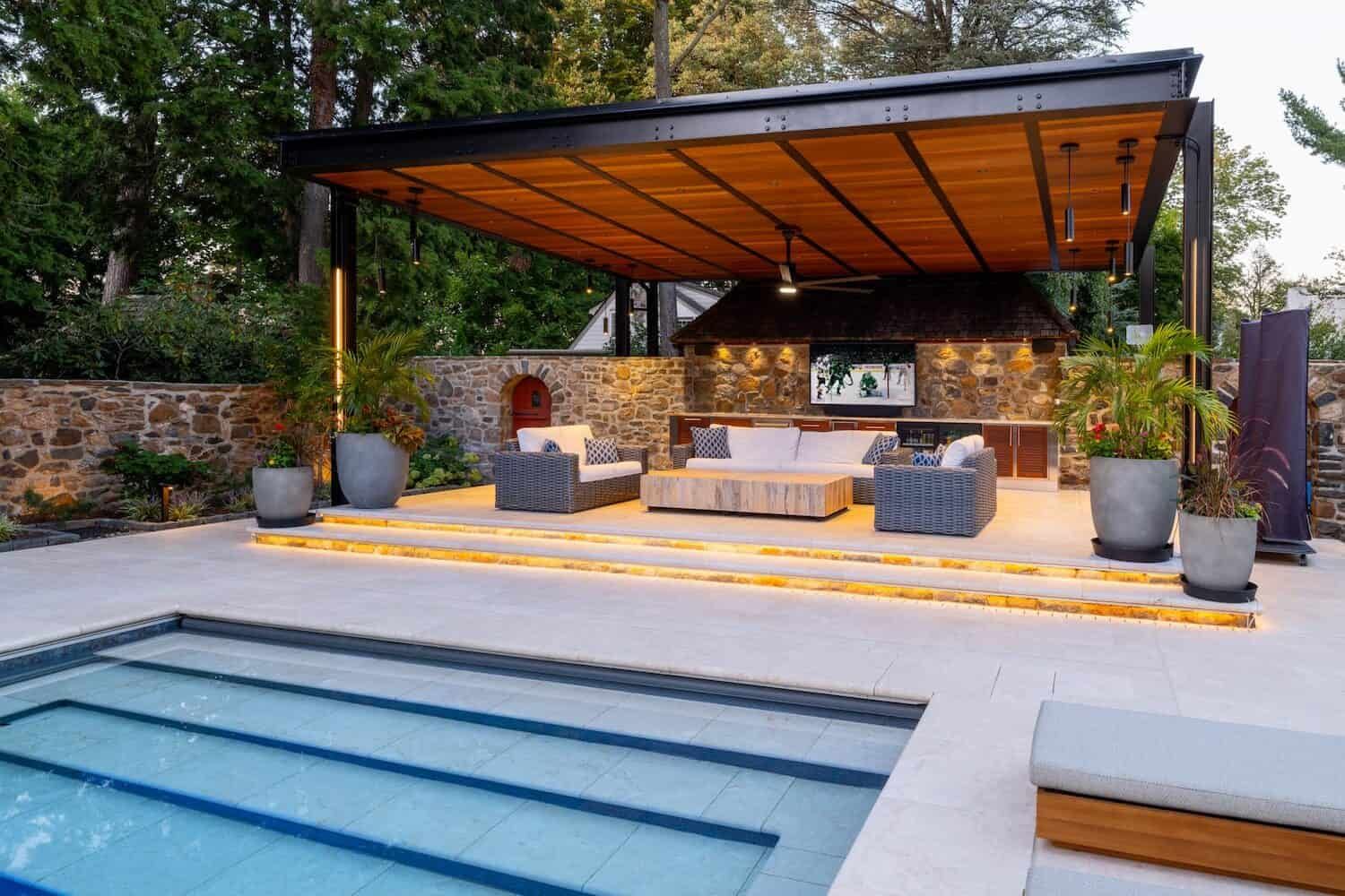 Modern backyard pool with covered lounge area, stone wall, and warm ambient lighting
