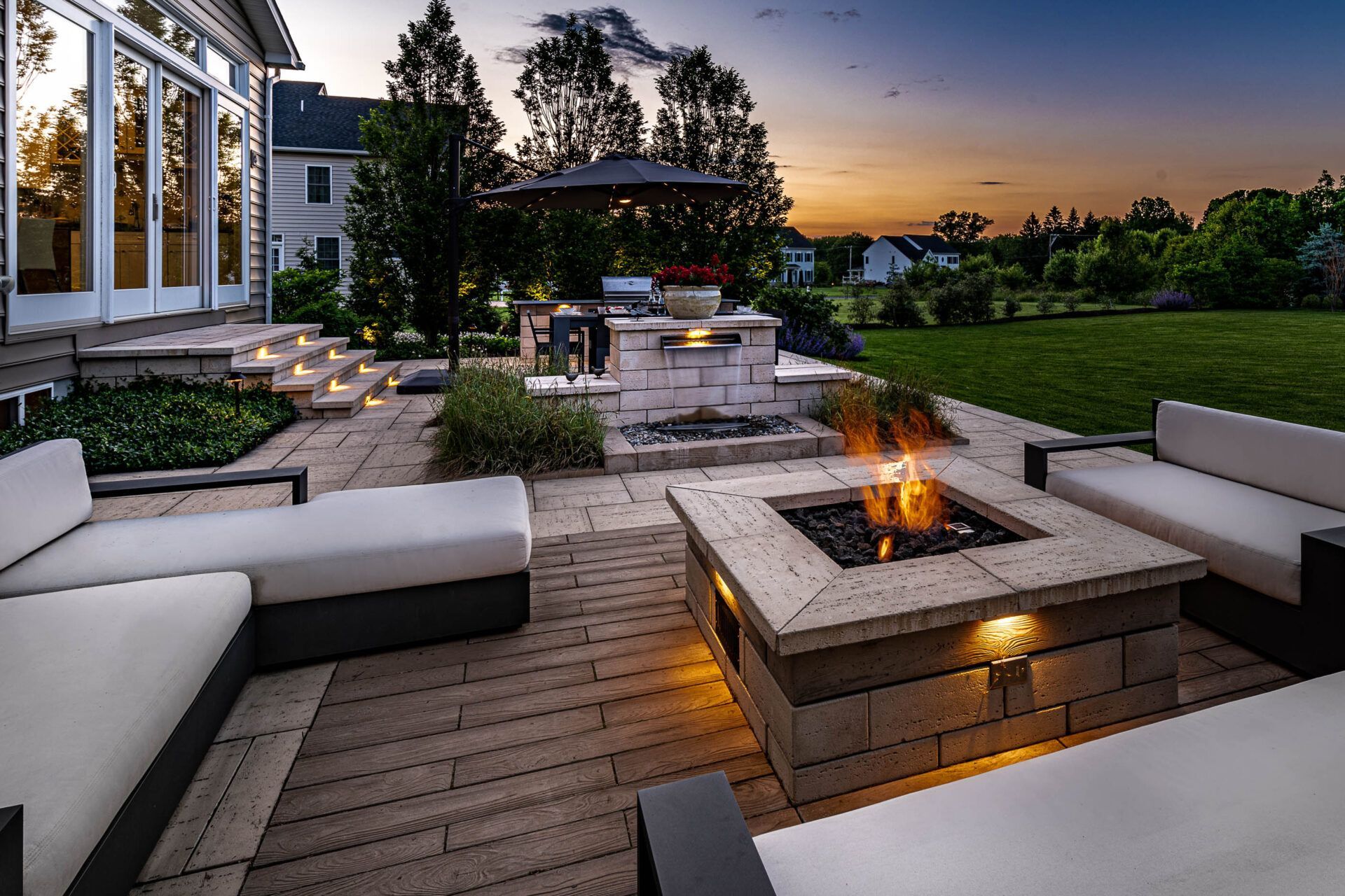 Modern patio at dusk with a lit fire pit, sectional seating, and landscaped backyard views