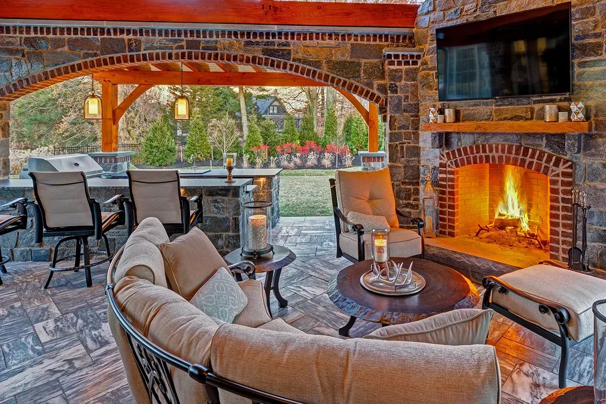 Covered patio with cushioned chairs, dining table, and lit brick fireplace overlooking a garden.