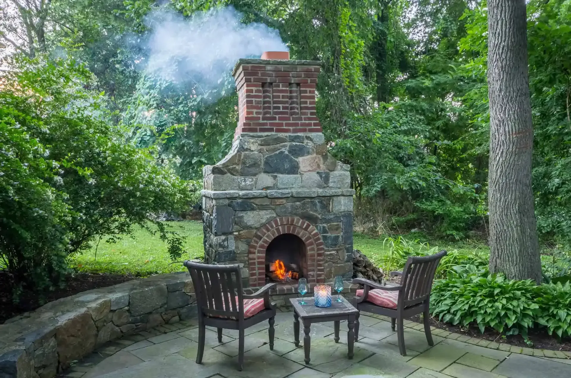 Stone outdoor fireplace with chairs and a small table in a leafy garden patio