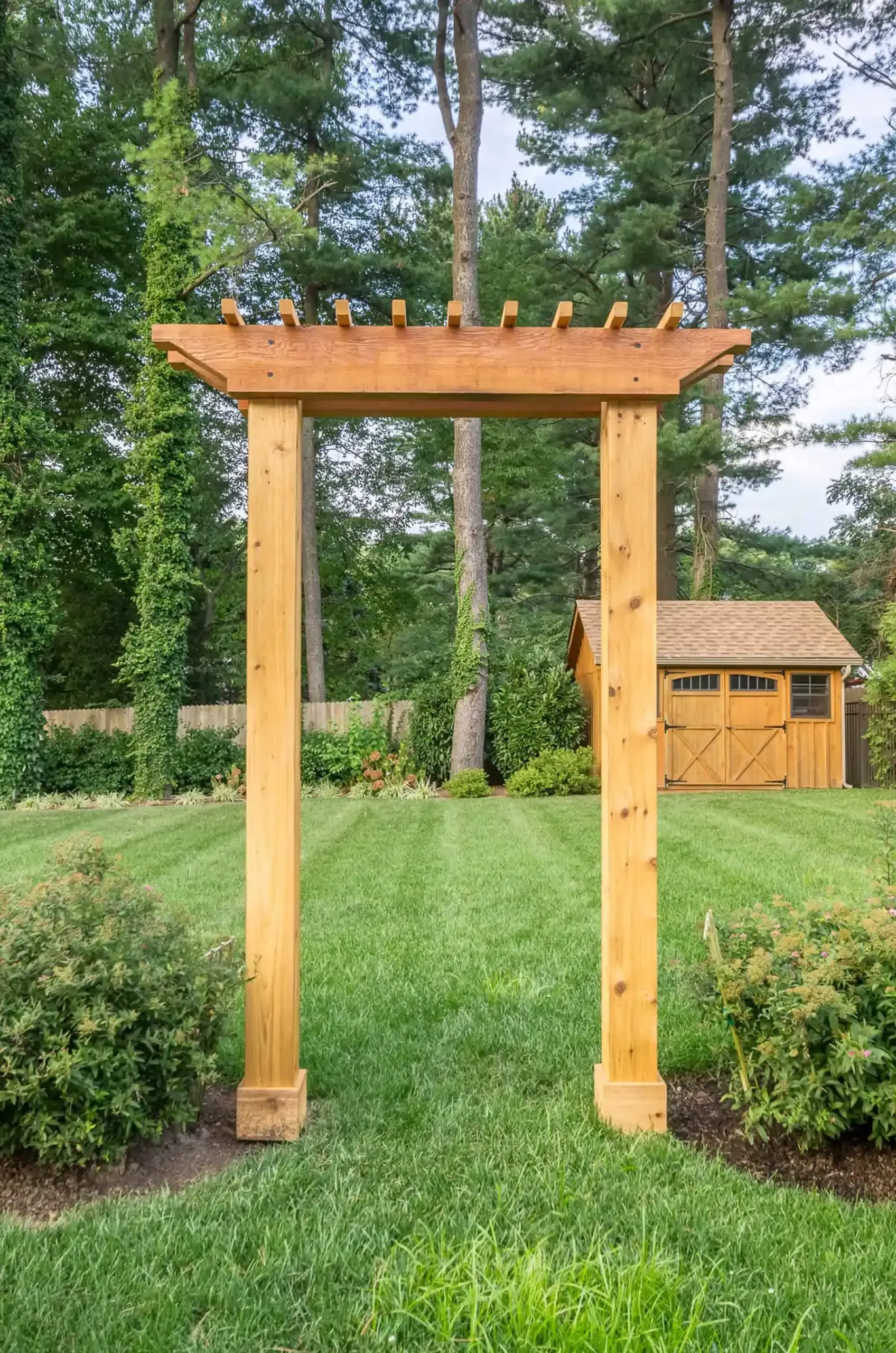 Wooden garden pergola arch over grass, with a small shed and trees in the background.