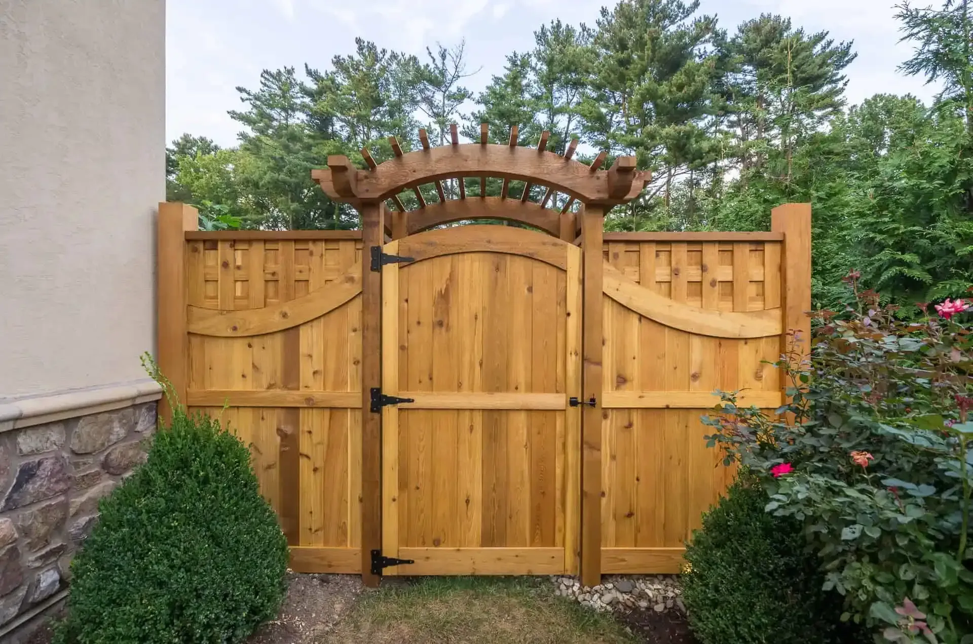 Wooden garden gate with arched top and lattice panels beside a stone house wall and shrubs