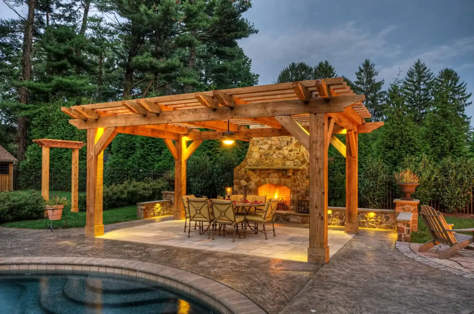 Wooden pergola with warm lights and seating beside a stone fireplace near a pool at dusk