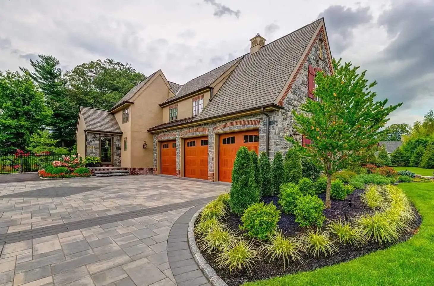 Stone house with orange garage doors, curved driveway, and landscaped garden under a cloudy sky