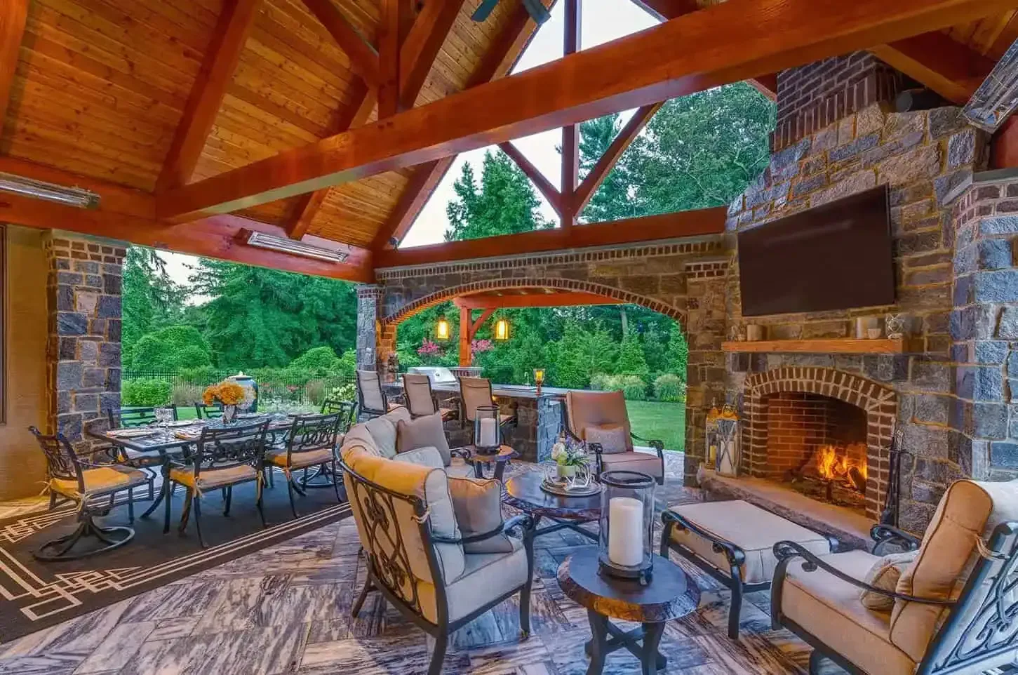 Stone patio lounge with wicker chairs and tables under a wooden pergola, next to a lit fireplace