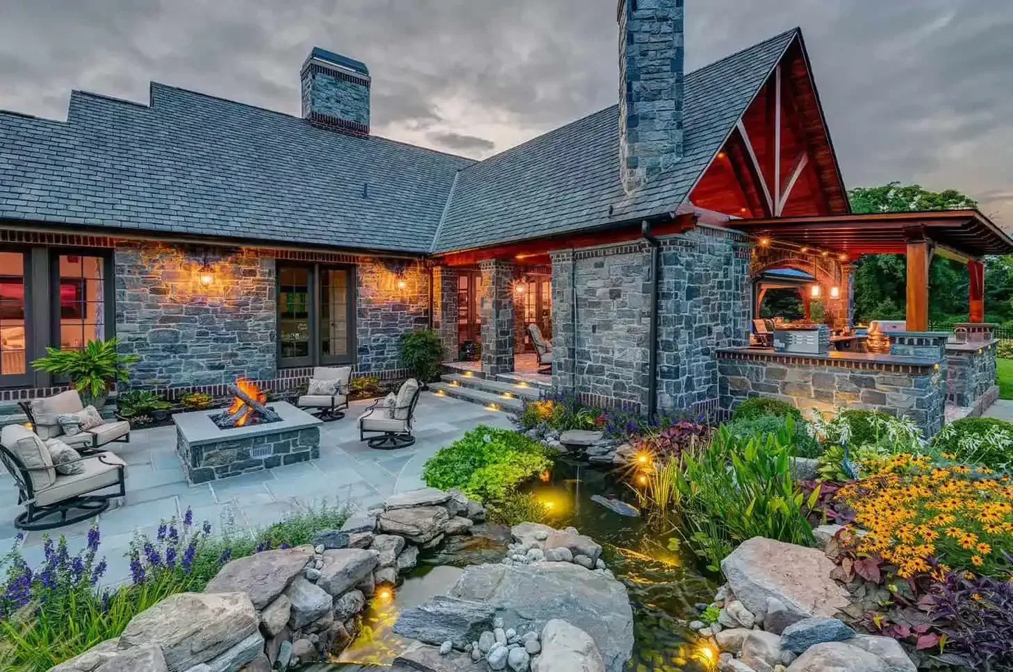 Stone house with lit patio, stone path, and colorful garden at dusk