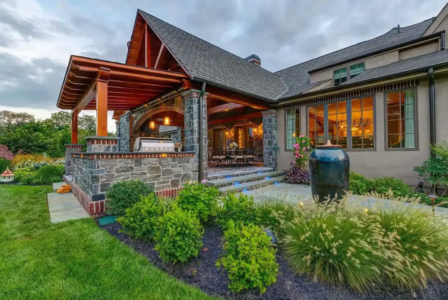 Modern house with a covered stone porch, warm lights, and landscaped garden at dusk