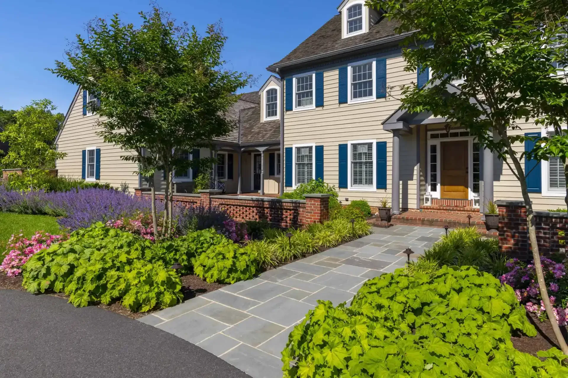 Landscaped suburban house with blue shutters, stone walkway, and blooming flowers