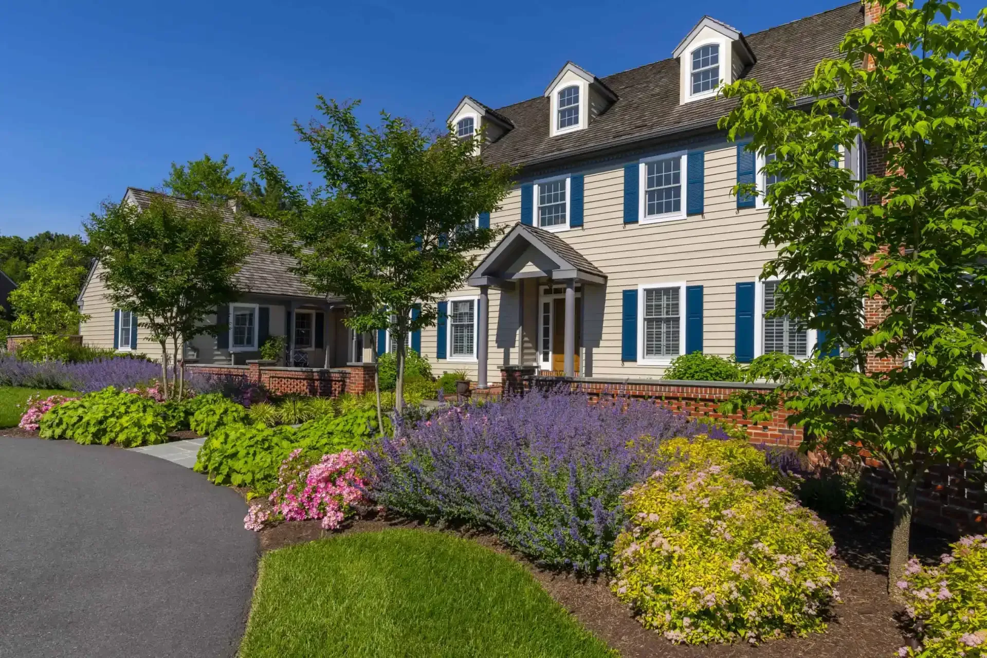 Large white colonial house with blue shutters, landscaped flower beds, and a driveway under a clear blue sky