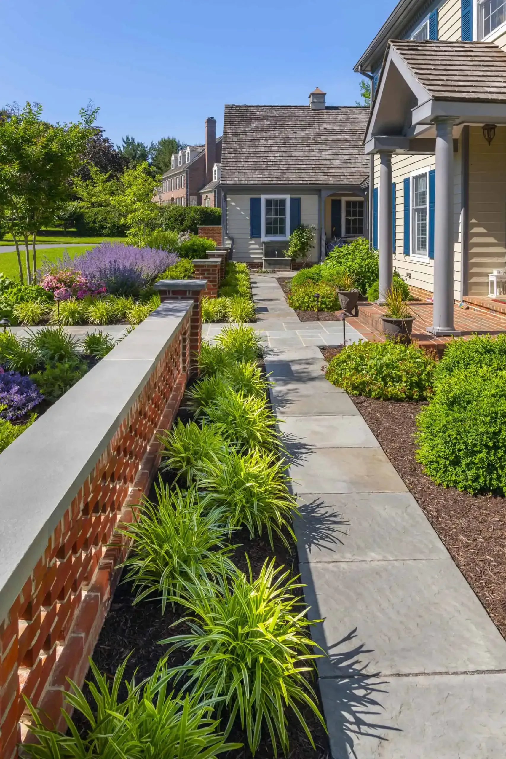 Landscaped front walkway beside a house, with stone path, brick wall, and purple flowers