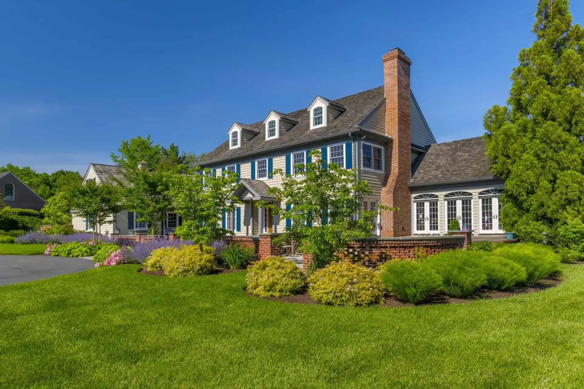 Large stone house with blue shutters, brick chimney, and a lush green lawn under a clear blue sky