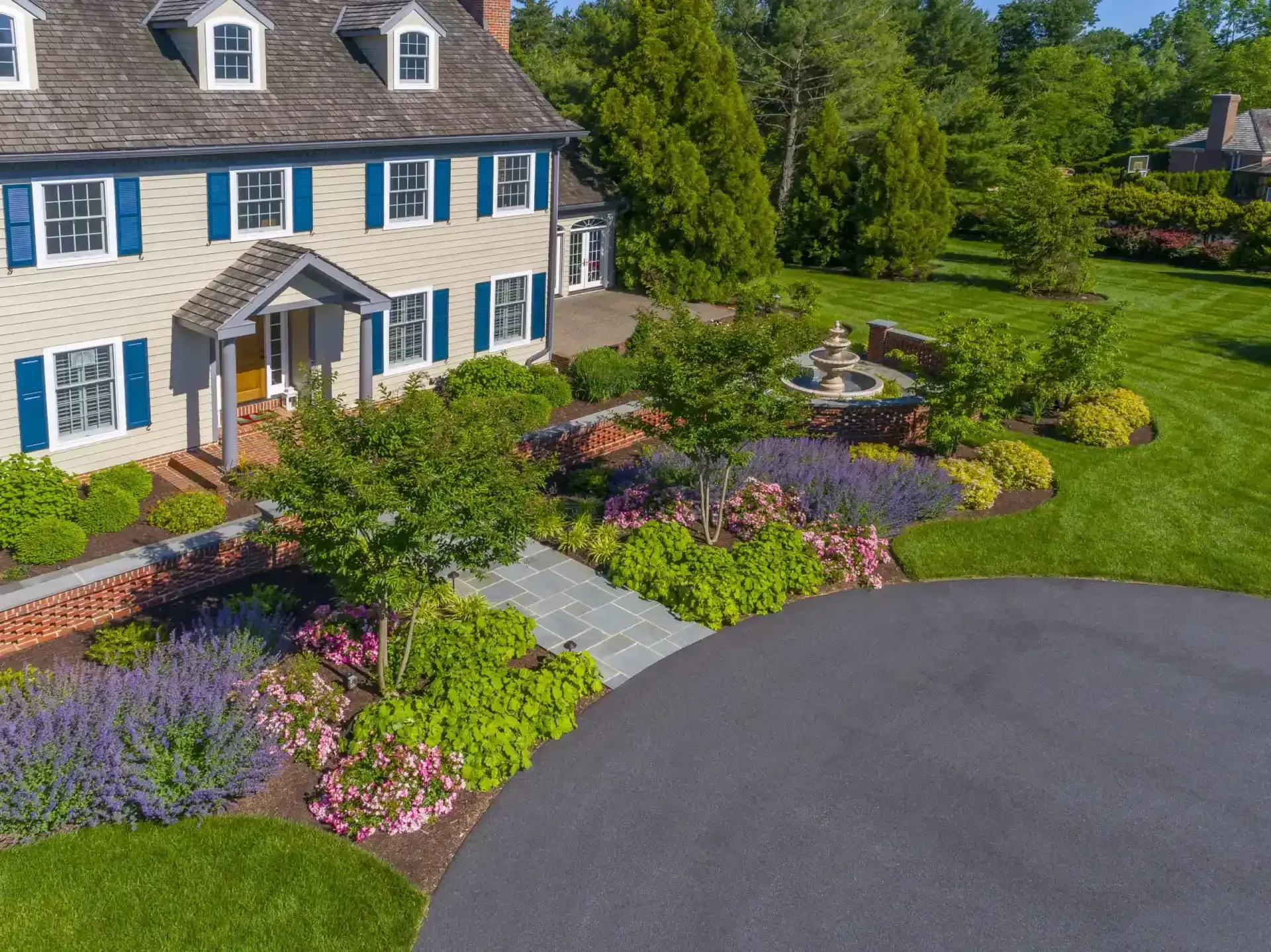 Stone house with blue shutters, colorful flower beds, and a curved driveway in front.