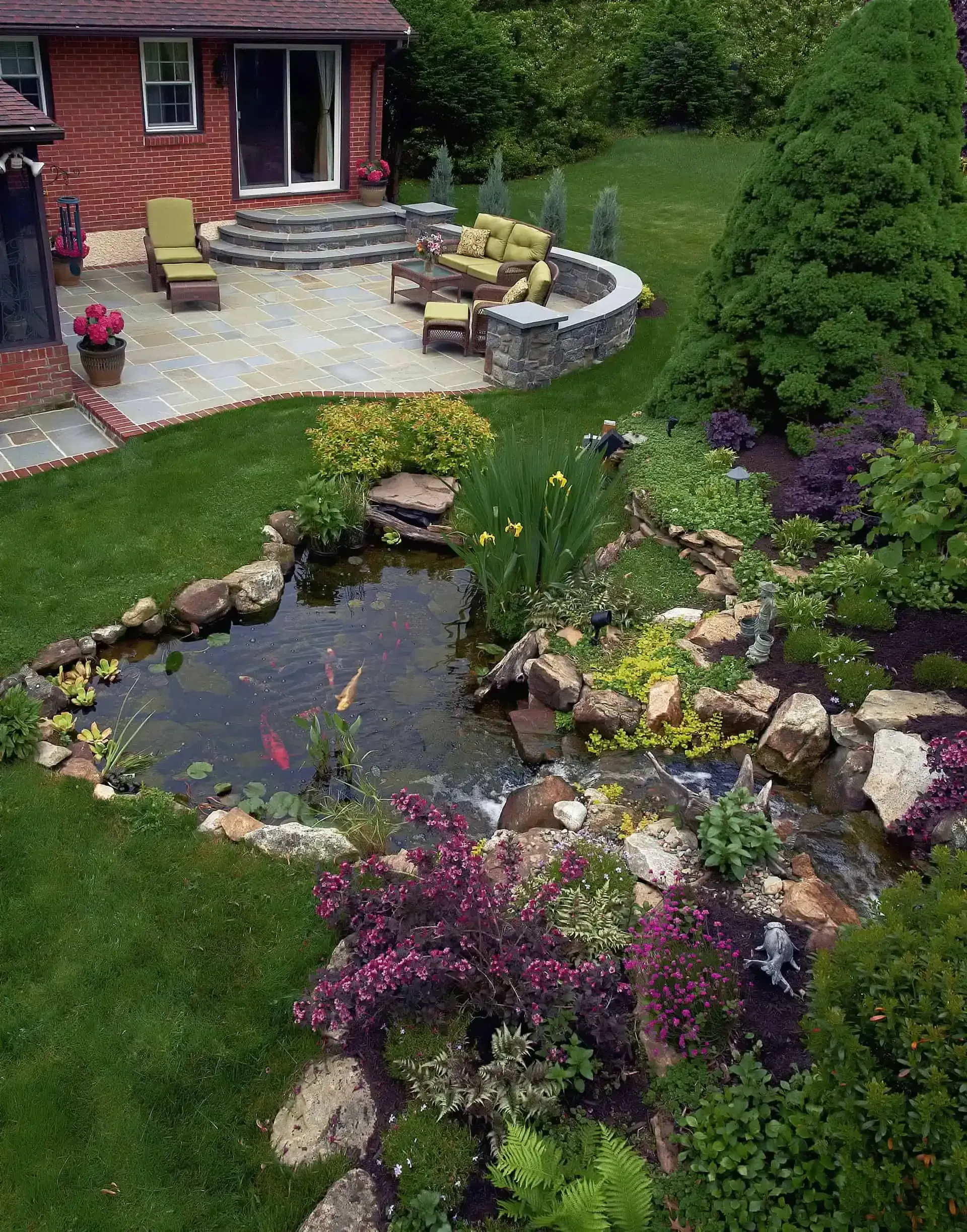 Backyard garden with a small pond, rocks, flowers, and a brick house patio nearby