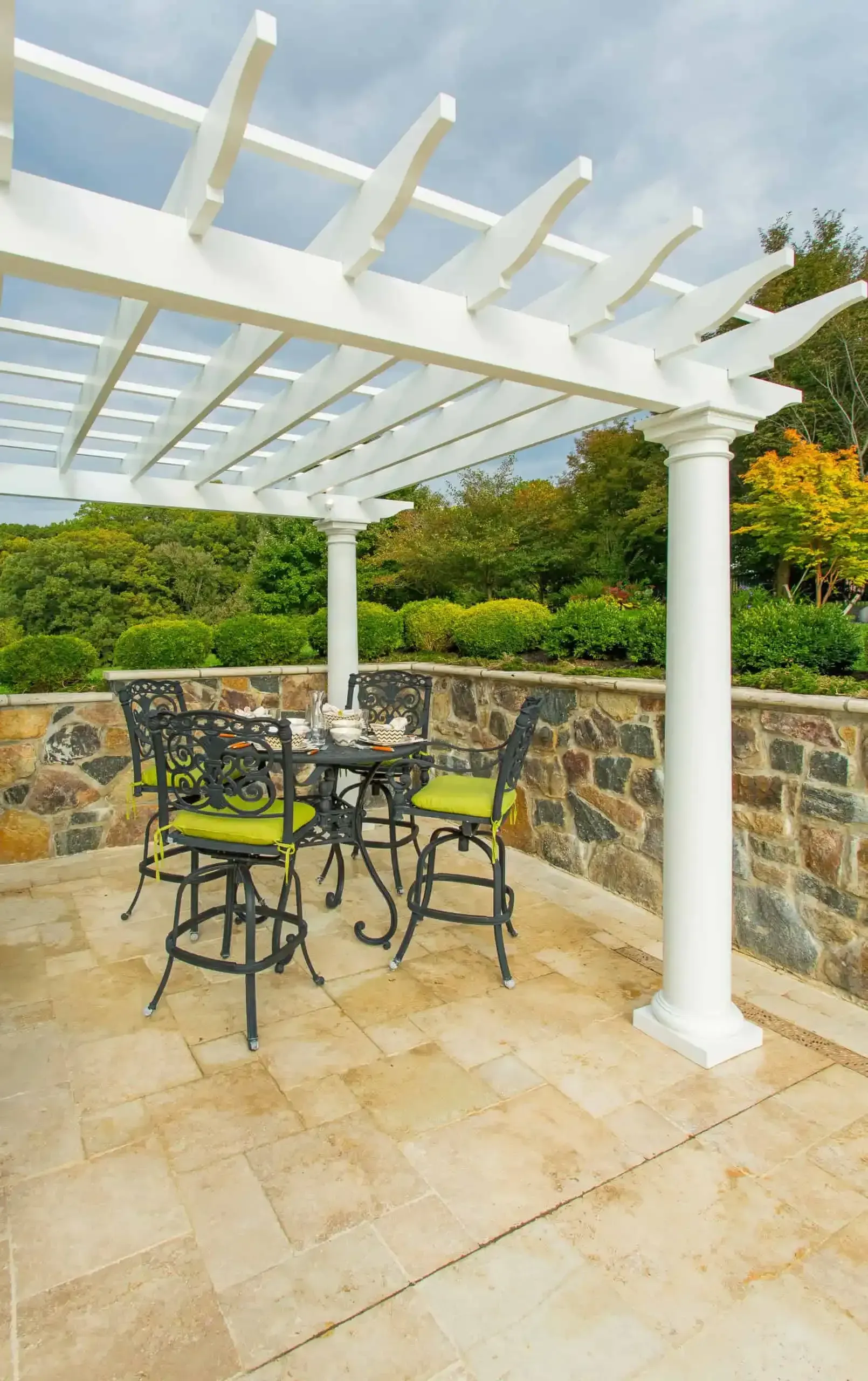 Patio with white pergola, black metal chairs, stone wall, and lush green garden view