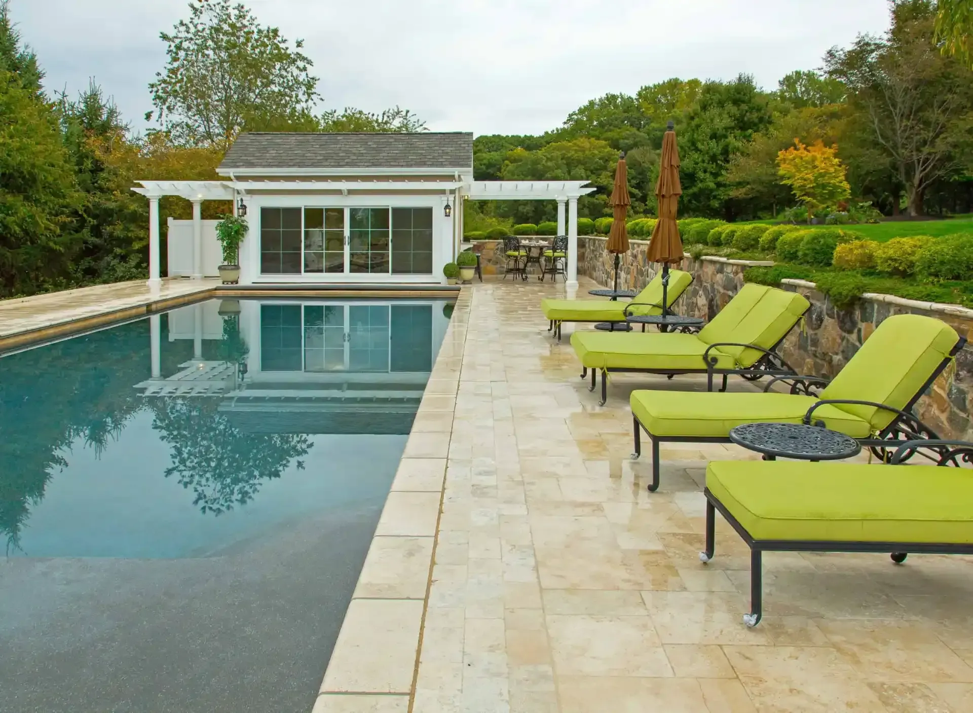 Backyard pool with green lounge chairs on a stone patio beside a modern cabana