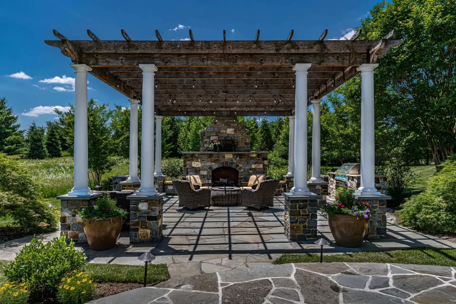 Stone patio pergola with white columns, wicker seating, potted plants, and a blue sky garden backdrop