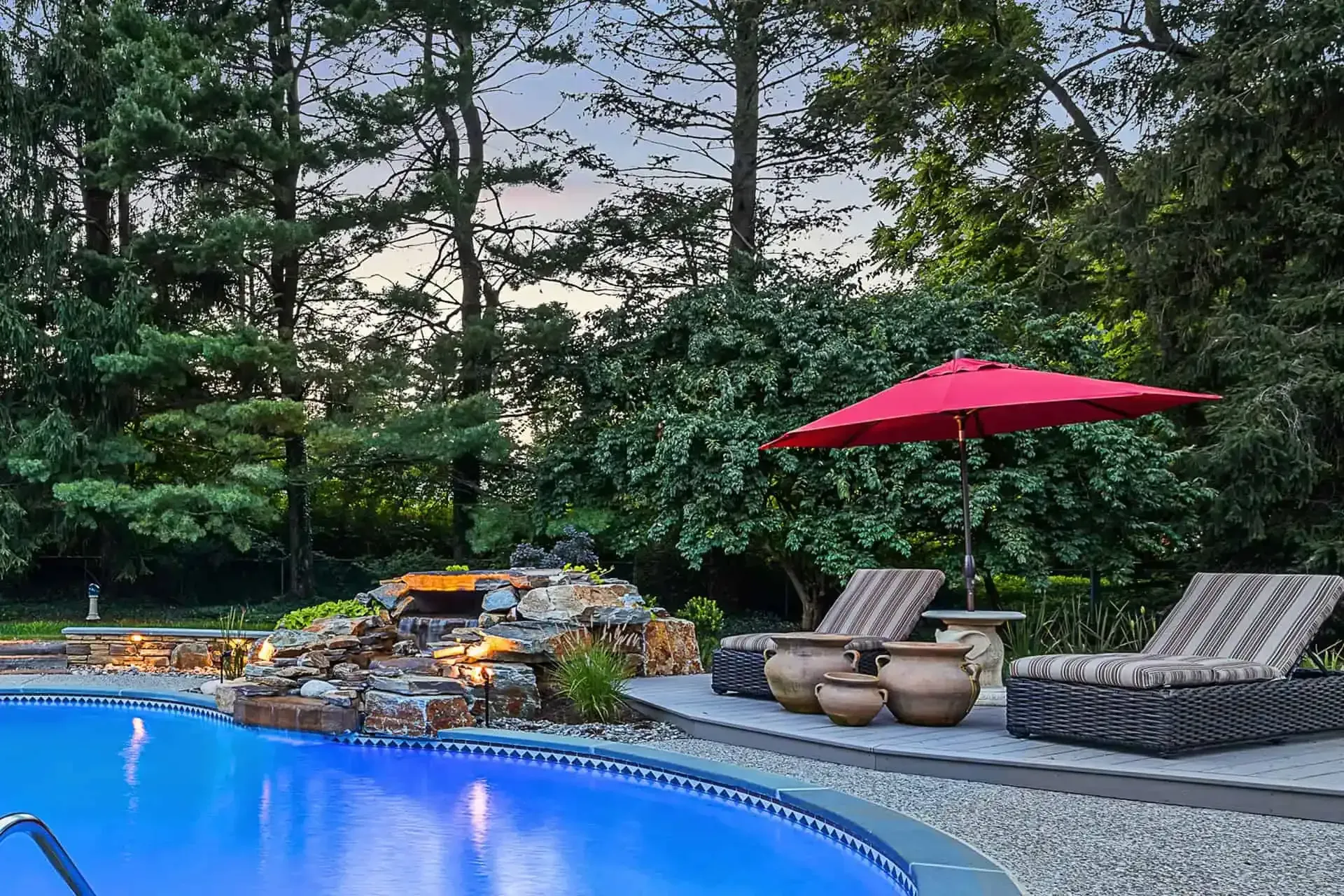 Blue pool beside lounge chairs, red umbrella, and lush trees in a landscaped backyard.