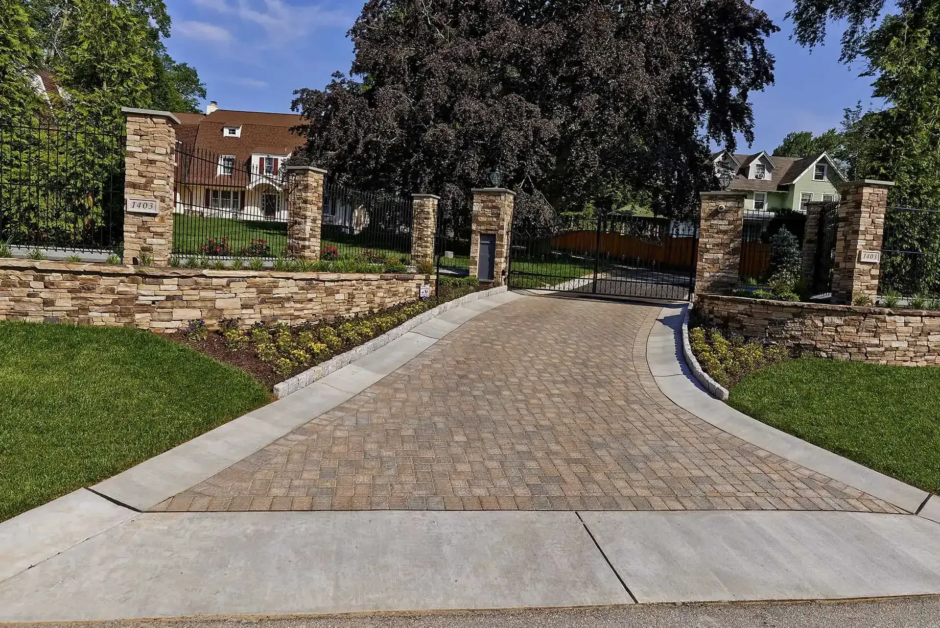 Brick driveway between stone walls leading to a house entrance on a sunny day