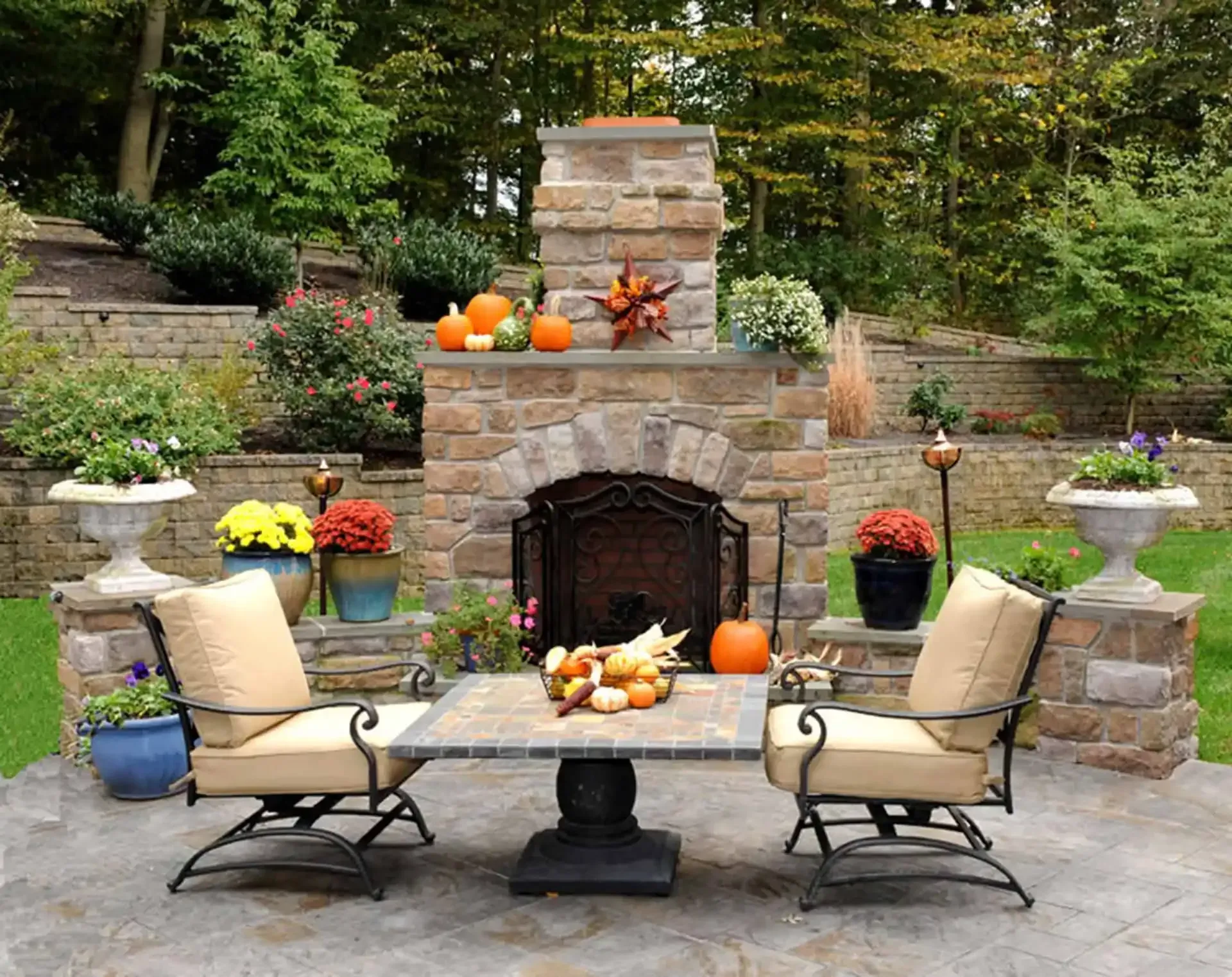 Fall patio with stone fireplace, pumpkins, chairs, and potted flowers in a garden setting