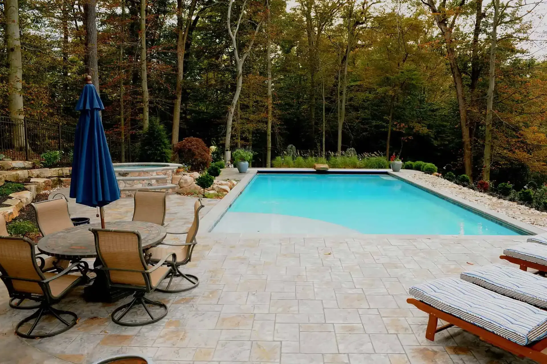 Backyard patio with a turquoise swimming pool, lounge chairs, and a closed blue umbrella amid trees