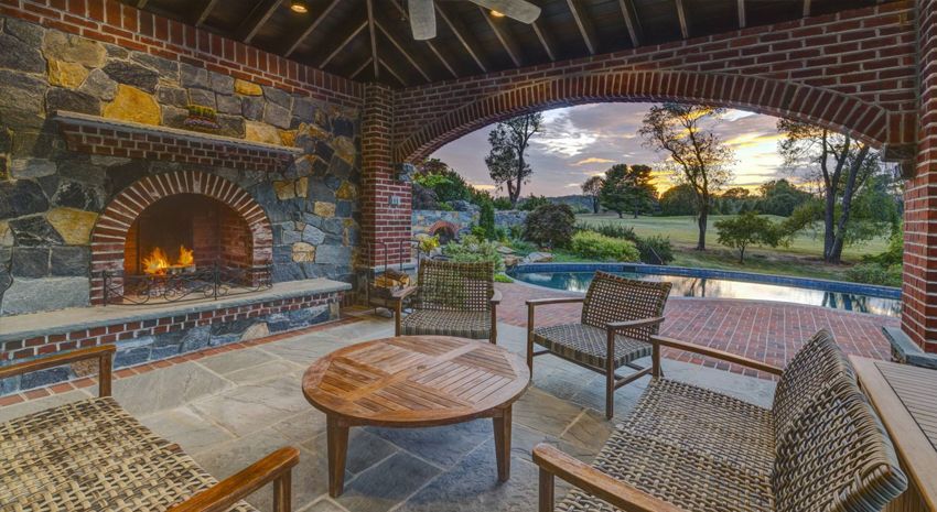 Covered patio with brick fireplace, wicker seating, and a pool view at sunset