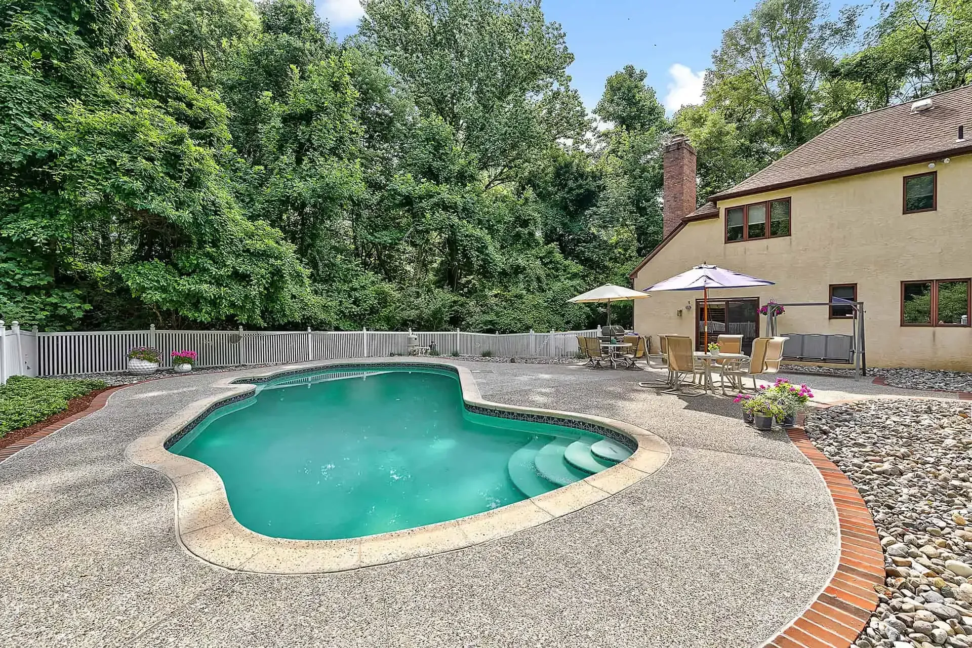 Backyard pool with turquoise water, stone patio, umbrellas, and a house surrounded by trees