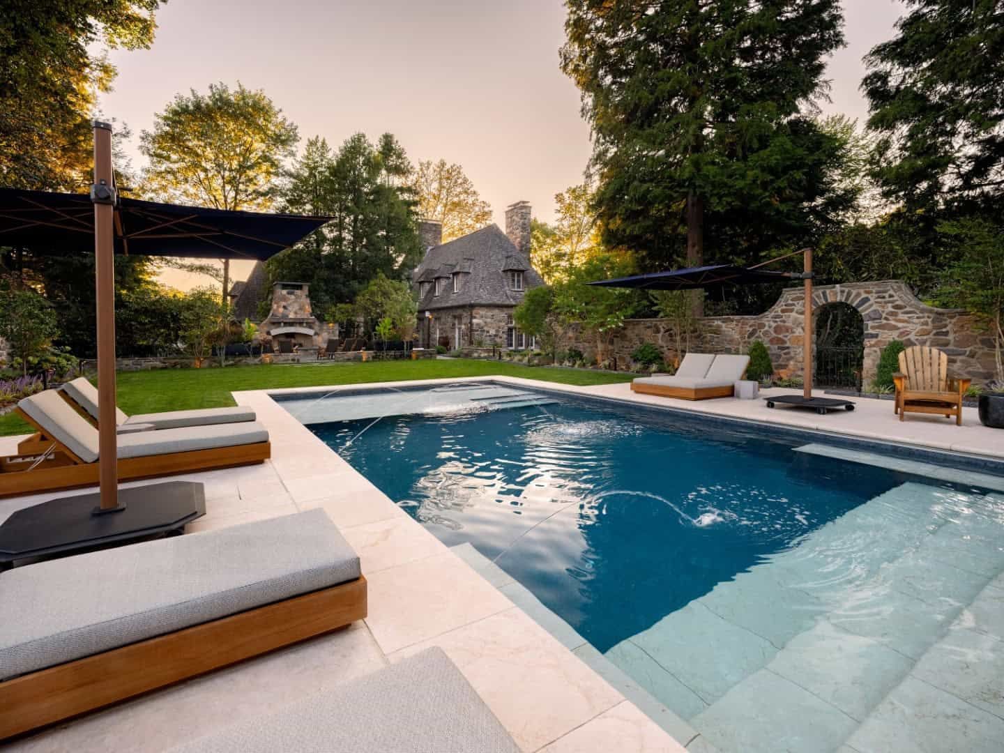 Modern backyard pool with lounge chairs, stone patio, and a rustic house in the background