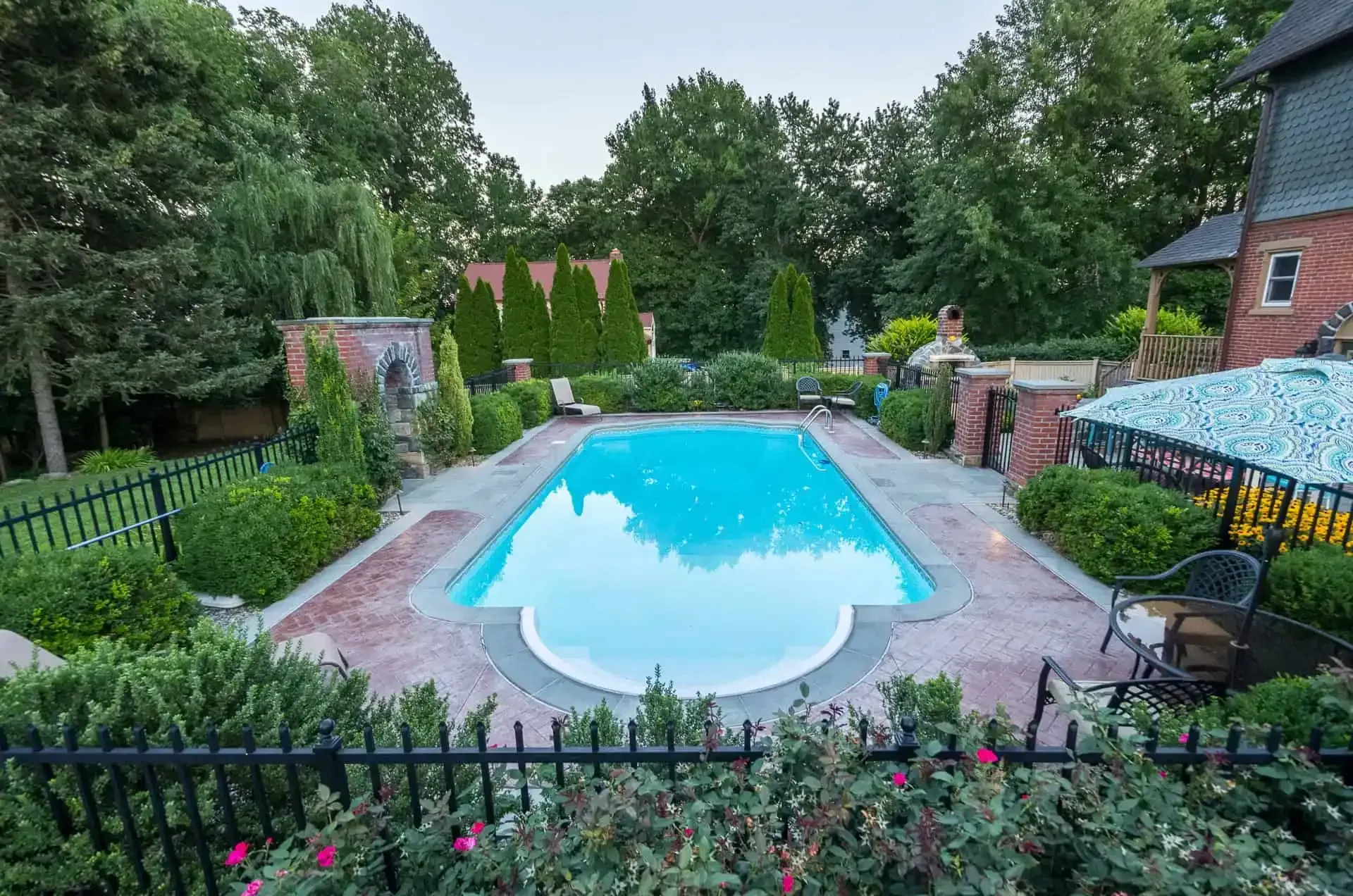 Backyard swimming pool surrounded by brick patio, fencing, and lush trees