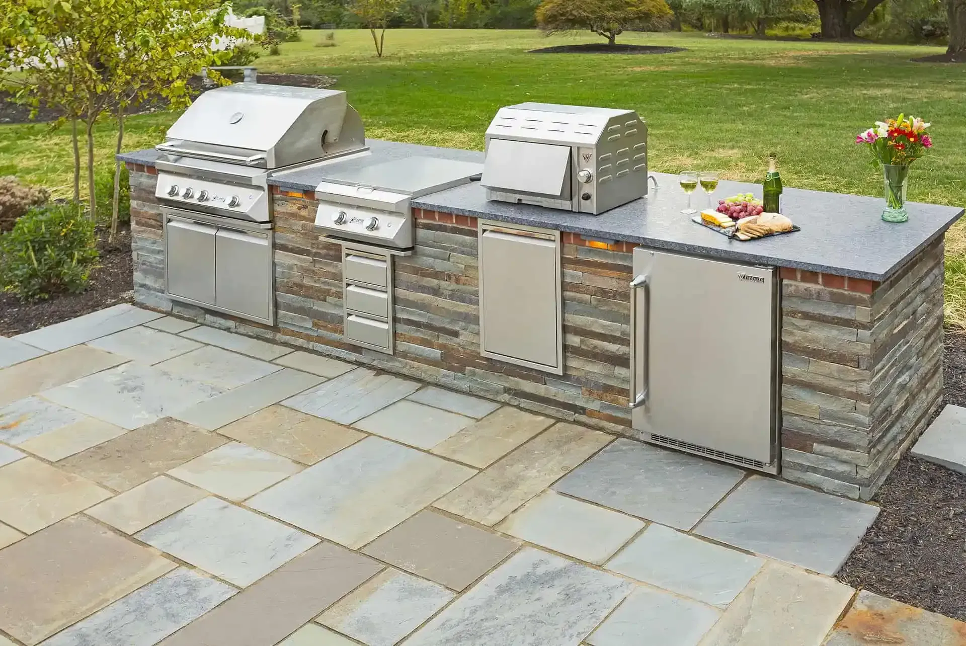Outdoor built-in stone barbecue island with grills, drawers, and side cabinet on a patio