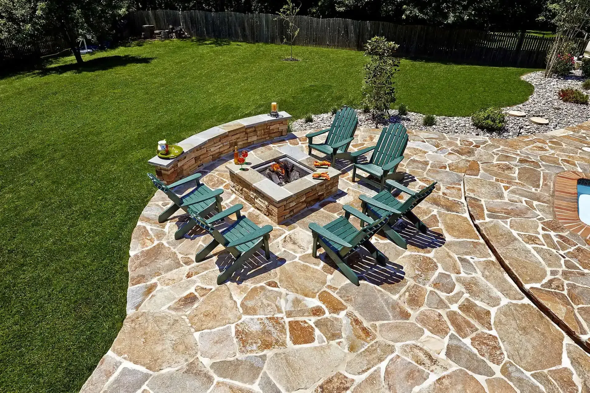 Stone patio with turquoise chairs around a fire pit beside a green lawn and trees