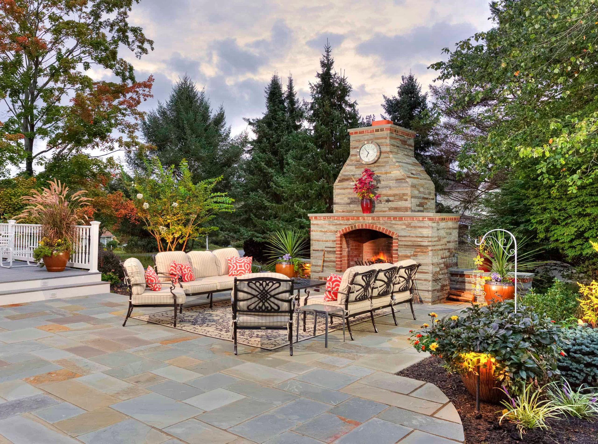 Stone patio with outdoor seating and a brick fireplace, surrounded by trees and string lights at dusk