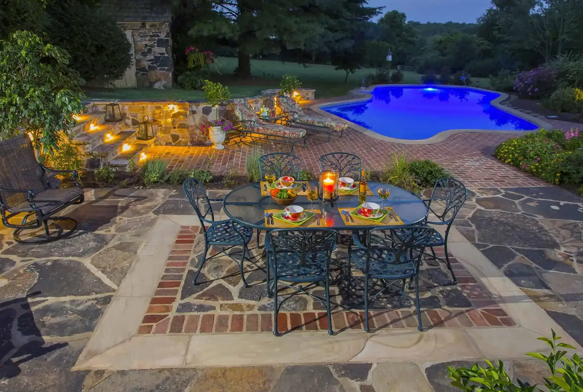 Stone patio dining area by a glowing backyard pool at dusk