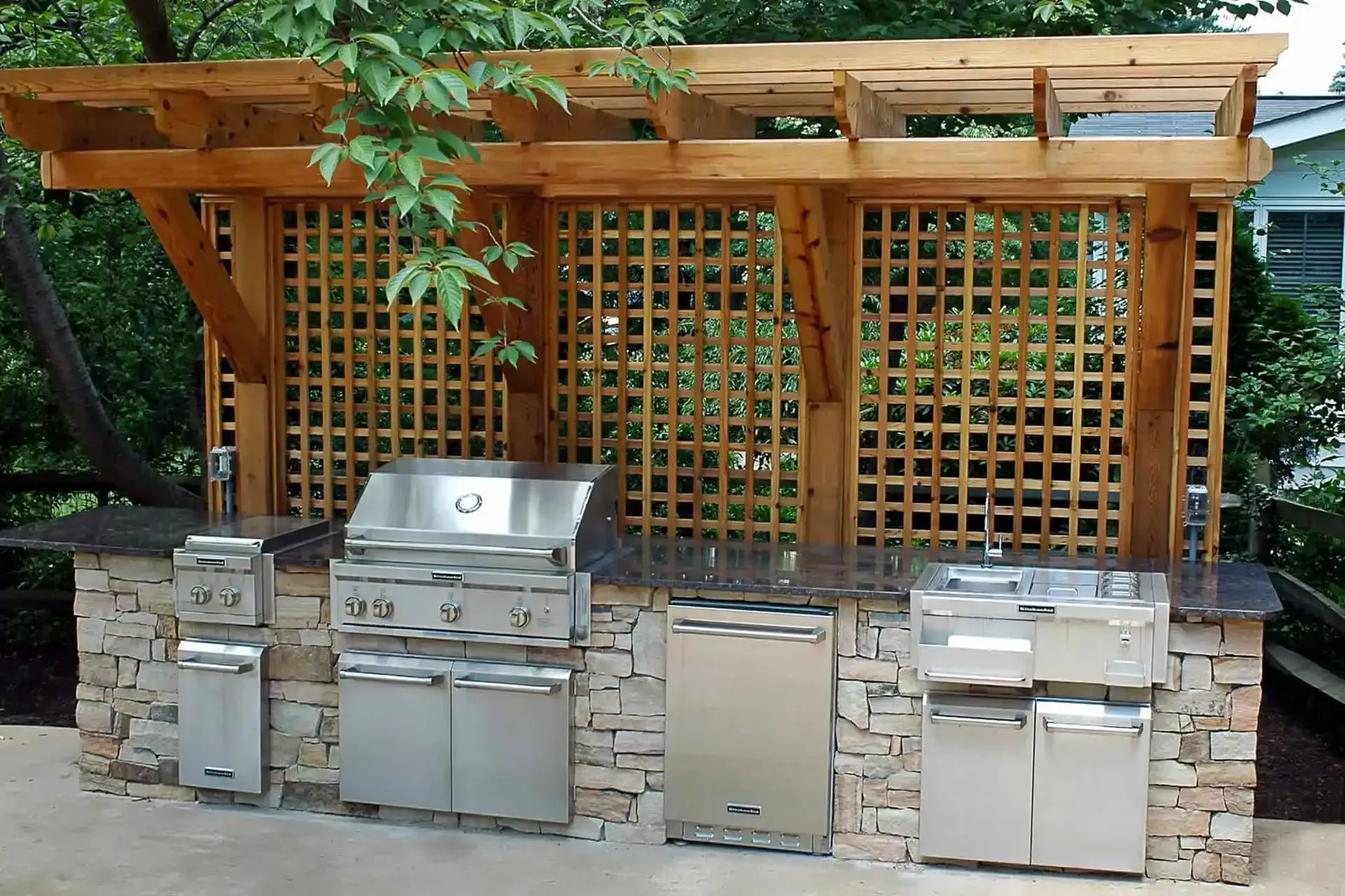 Outdoor stone grill station with stainless steel grills and a wooden pergola against a lattice wall