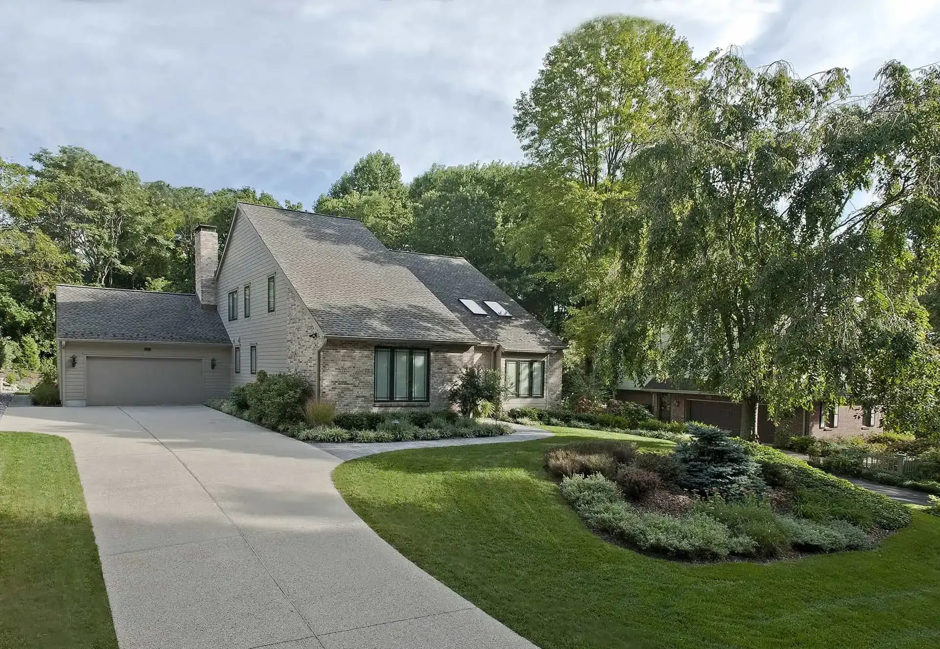 Stone house with attached garage, curved driveway, and landscaped front yard under tall trees