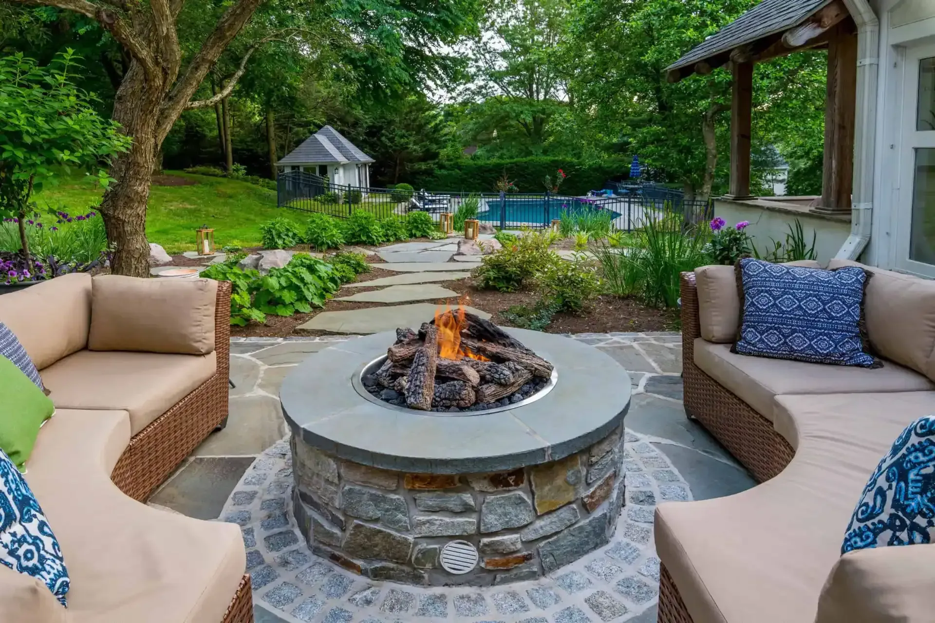 Backyard patio with fire pit, cushioned chairs, stone path, lush garden, and pool in the background