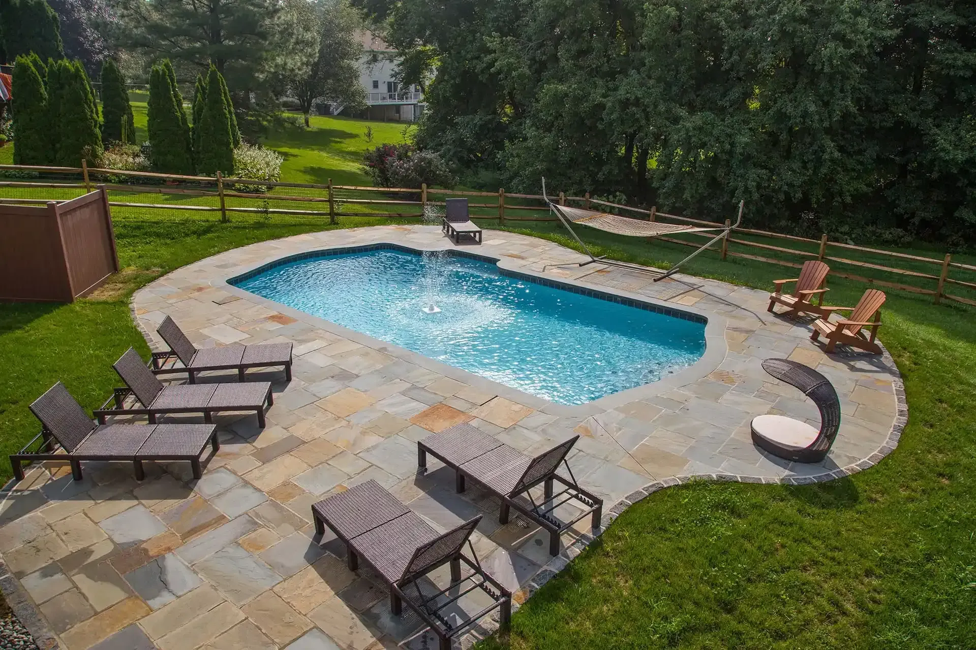 Backyard swimming pool with stone patio, lounge chairs, and green lawn surrounded by trees